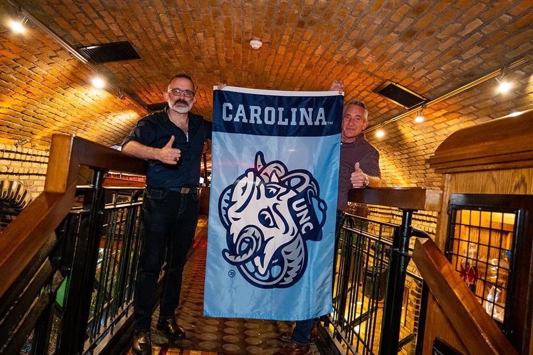 Two men standing inside O'Connell's Bar Dublin with a Carolina Gunc flag.